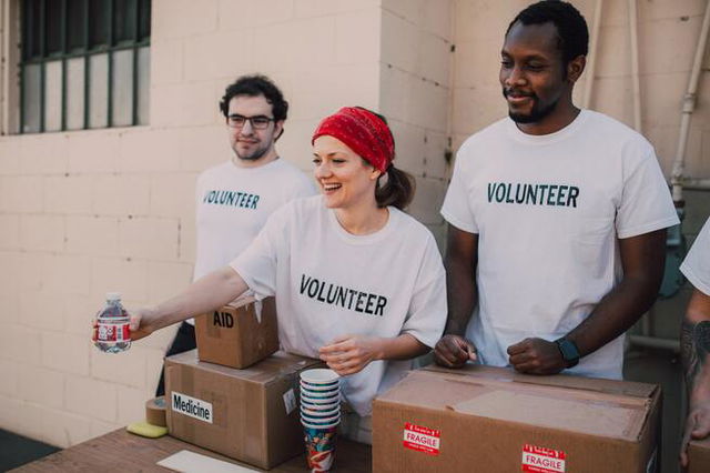 three people wearing teeshirts that say volunteers standing behind boxes handing out bottles, a female volunteer in a bandana is smiling.